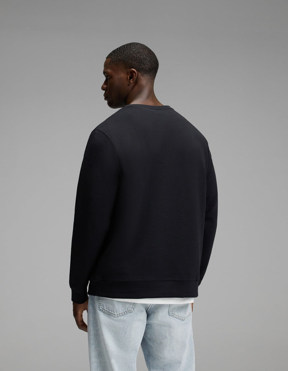 A man stands facing away, wearing the Cozy Earth Men’s StretchTech Crewneck in black and light blue jeans against a plain gray background.