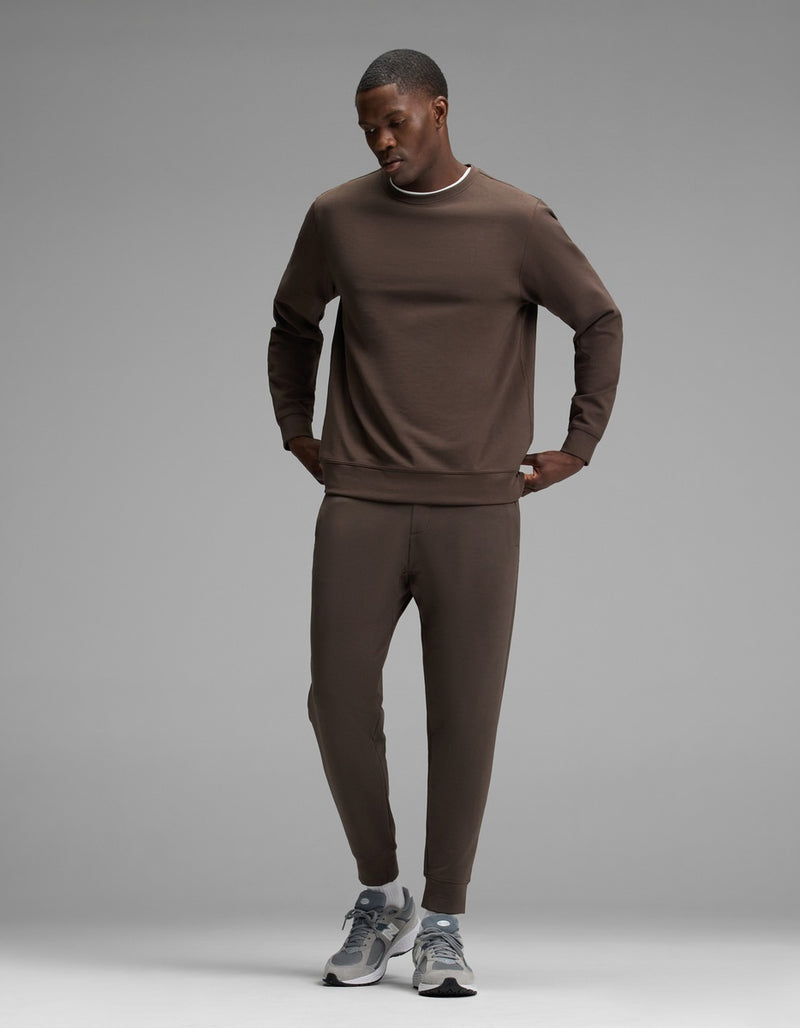 A man stands against a plain gray background, wearing the Cozy Earth Men’s StretchTech Crewneck & Jogger Set in brown, paired with gray running shoes. He looks down, hands in his pockets.