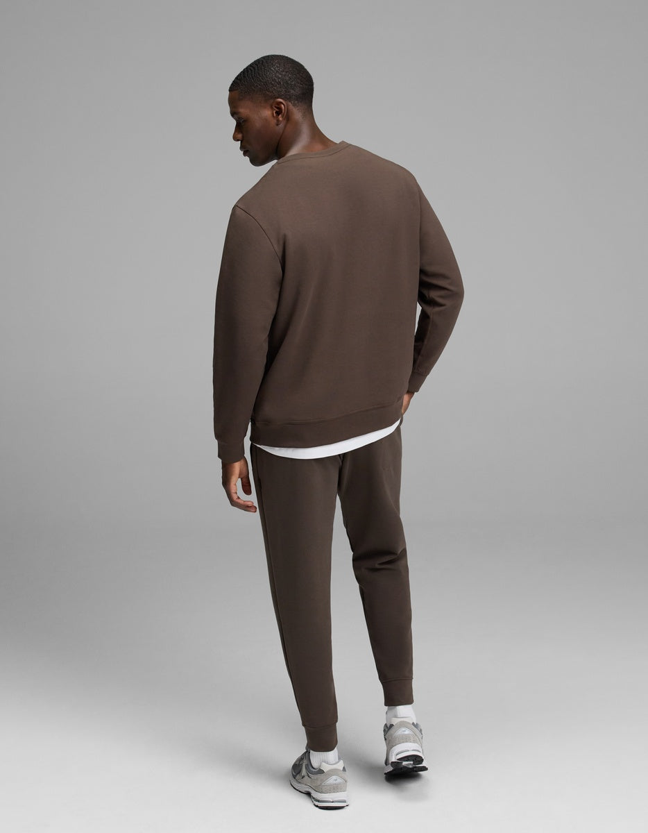 A man stands facing away, wearing Cozy Earth’s Men’s StretchTech Crewneck & Jogger Set in brown over a white shirt, paired with gray sneakers, against a plain gray background.|Color:Cacao