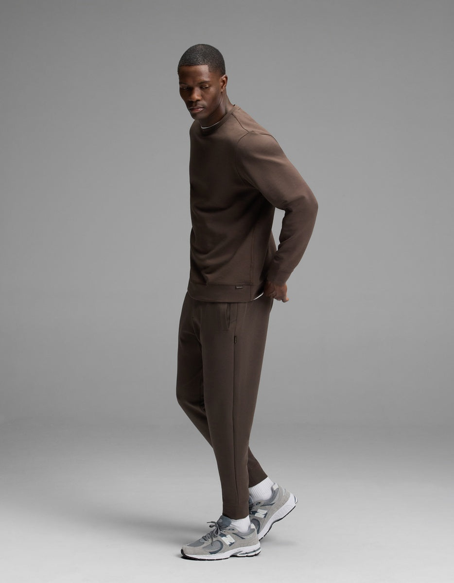 A man wearing the Cozy Earth Men’s StretchTech Crewneck & Jogger Set in brown stands sideways on a gray background, looking toward the camera. He completes his look with gray and white sneakers.
