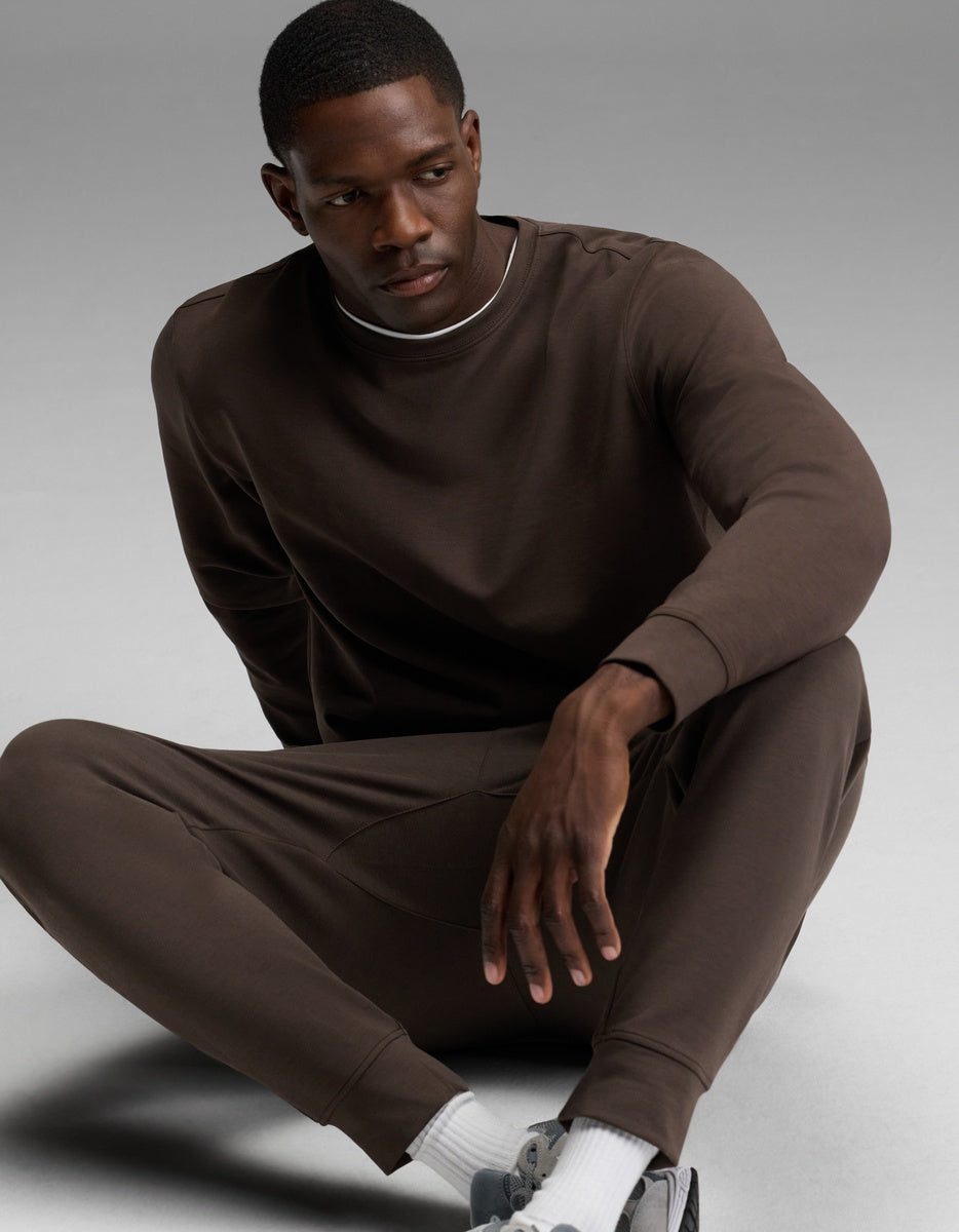 A man sits on the ground in a relaxed pose, wearing the Cozy Earth Men’s StretchTech Crewneck & Jogger Set in brown, paired with white socks and gray sneakers, against a plain gray background.