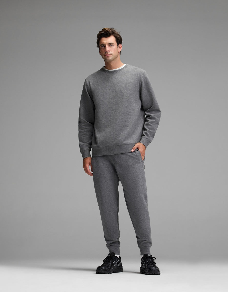 A man stands facing forward, wearing the Cozy Earth Men’s StretchTech Crewneck & Jogger Set in gray with black sneakers, set against a plain gray background.