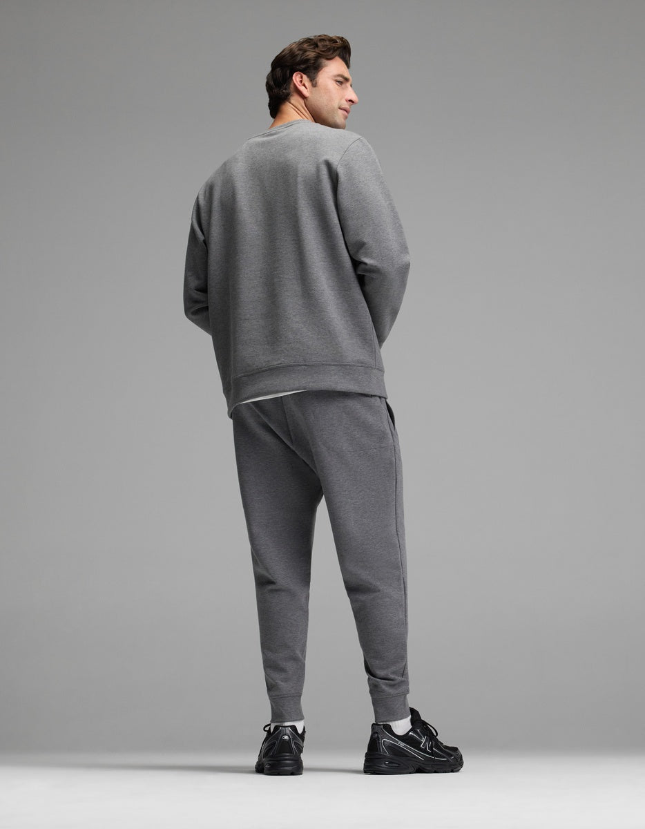 A person stands facing away, wearing the Cozy Earth Men’s StretchTech Crewneck & Jogger Set in grey with black sneakers, against a plain grey background.