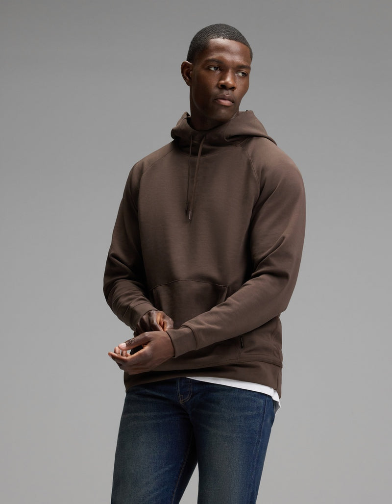 A man wearing the Cozy Earth Men’s StretchTech Hoodie and blue jeans stands against a plain gray background, looking to his left with his hands loosely clasped in front of him.