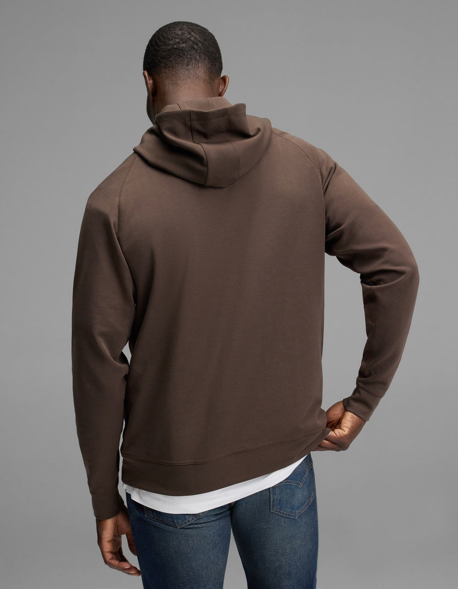 A person wearing the Cozy Earth Men’s StretchTech Hoodie in brown and blue jeans stands with their back to the camera against a plain gray background.