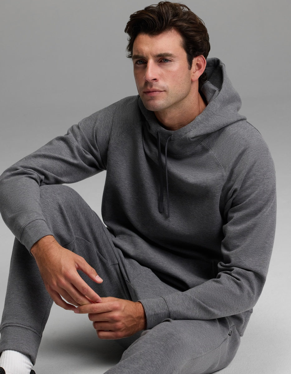 A man wearing the Cozy Earth Men’s StretchTech Hoodie and matching sweatpants sits on the floor against a plain gray background, looking slightly to the side with a neutral expression.