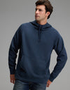 A man wearing the Cozy Earth Men’s StretchTech Hoodie in navy blue and jeans stands with one hand in his pocket against a plain gray background.|Color:Eclipse