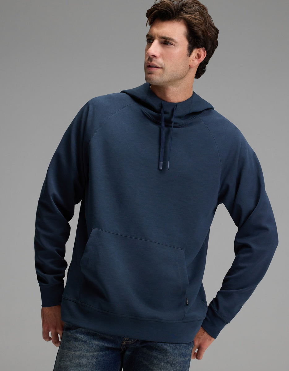 A man wearing the Cozy Earth Men’s StretchTech Hoodie in navy blue and jeans stands with one hand in his pocket against a plain gray background.|Color:Eclipse