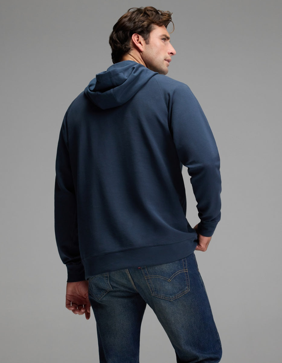 A man wearing the Cozy Earth Men’s StretchTech Hoodie in blue and a pair of jeans stands with his back to the camera, head turned slightly to the side, set against a plain gray background.