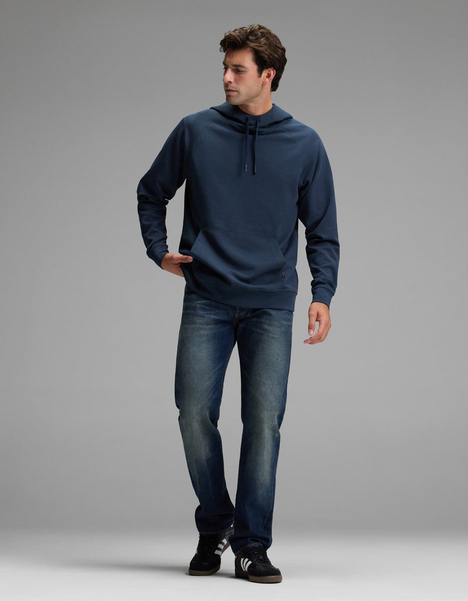 A man wearing the Cozy Earth Men’s StretchTech Hoodie, blue jeans, and black sneakers walks against a light gray background, looking to his right with one hand in his pocket.
