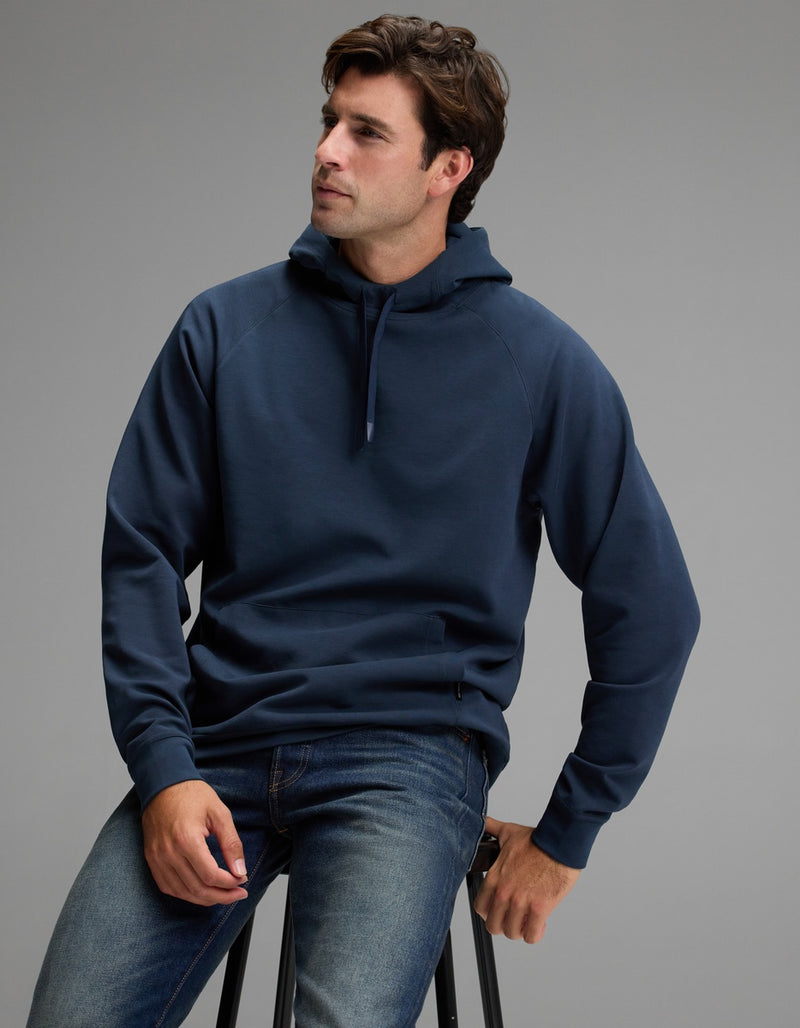 A man wearing the Cozy Earth Men’s StretchTech Hoodie in dark blue and jeans sits on a stool against a plain gray background, looking slightly to the side.