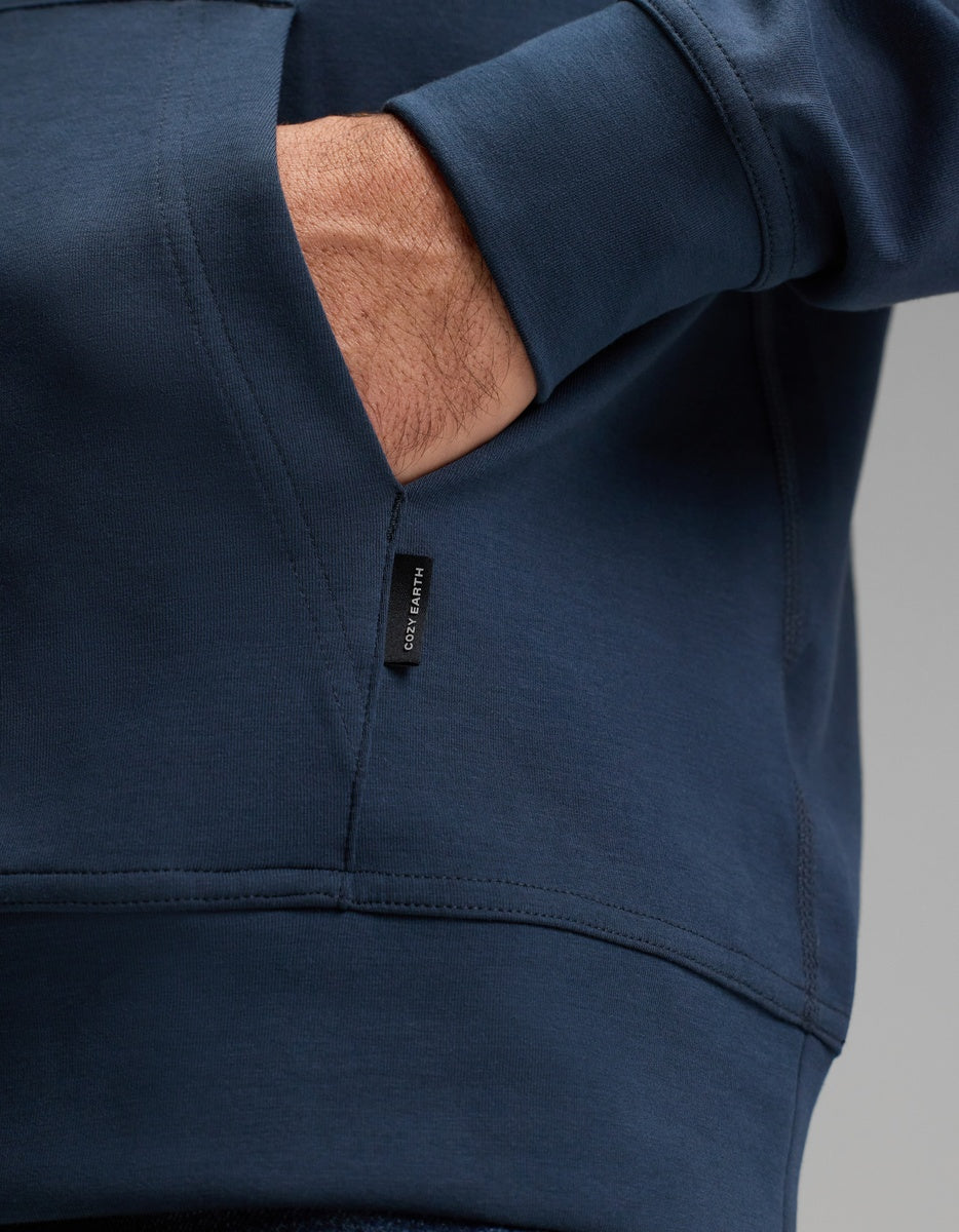 A close-up of a person’s hand partially tucked into the pocket of a dark blue Cozy Earth Men’s StretchTech Hoodie, featuring a small black fabric tag on the seam.