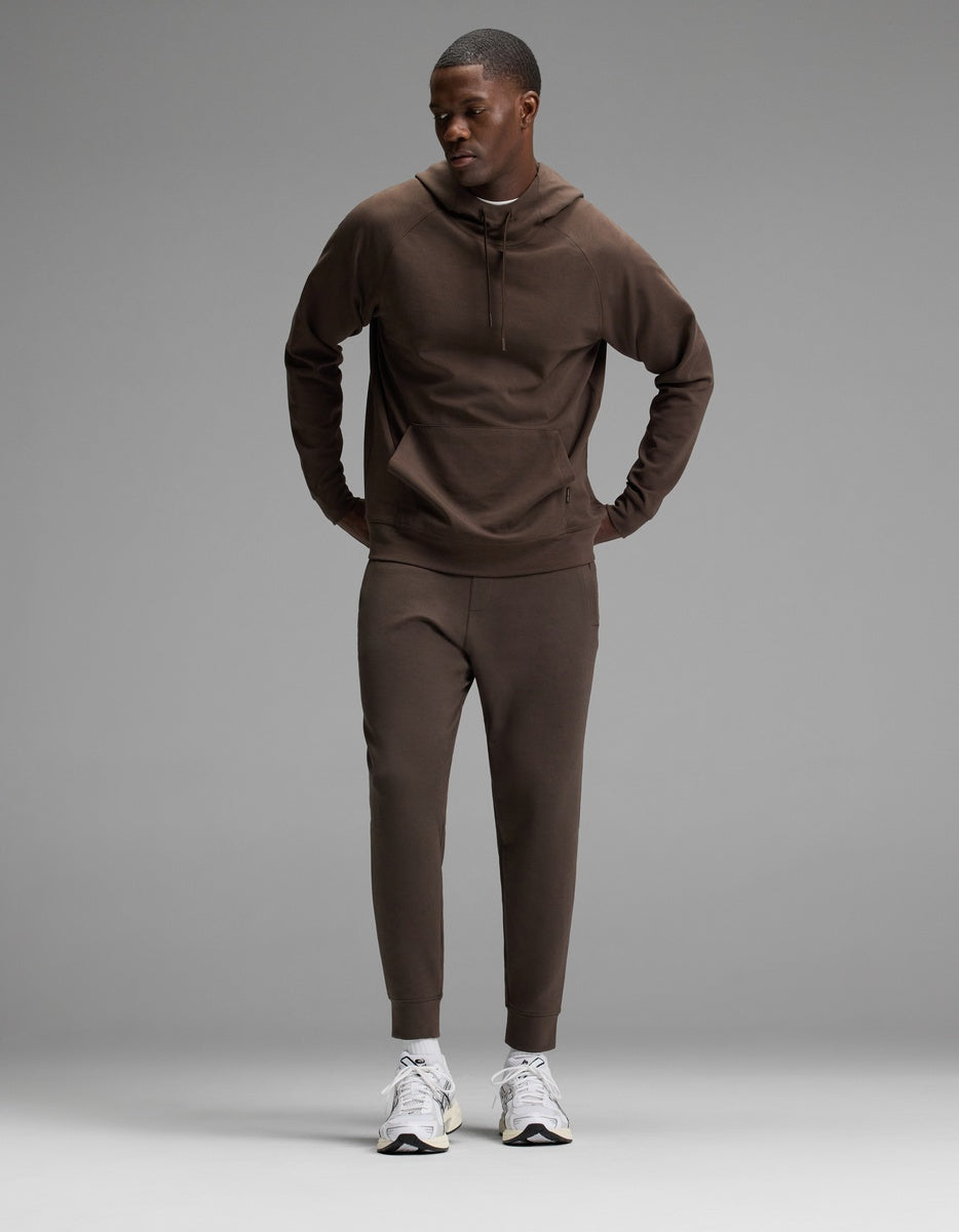 A man stands against a plain gray background, wearing the Cozy Earth Men’s StretchTech Hoodie & Jogger Set in brown with white sneakers, looking slightly down and to the side with his hands in his pockets.|Color:Cacao