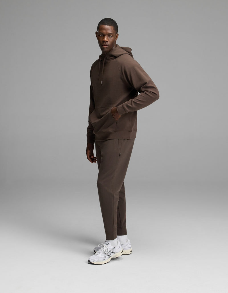 A man wears the Cozy Earth Men’s StretchTech Hoodie & Jogger Set in brown, standing in a neutral pose against a plain gray background and paired with white sneakers.