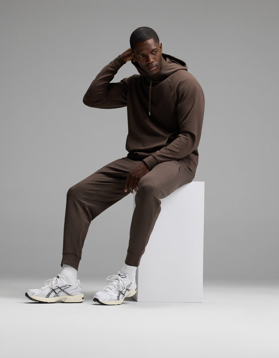 A man poses in a studio with a neutral background, sitting on a white block and wearing the Cozy Earth Men’s StretchTech Hoodie & Jogger Set paired with white sneakers and socks.