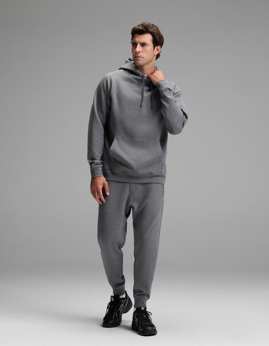 A man wearing the Cozy Earth Men’s StretchTech Hoodie & Jogger Set in gray stands against a plain gray background, looking to the side with one hand touching his hoodie, and wearing black sneakers.