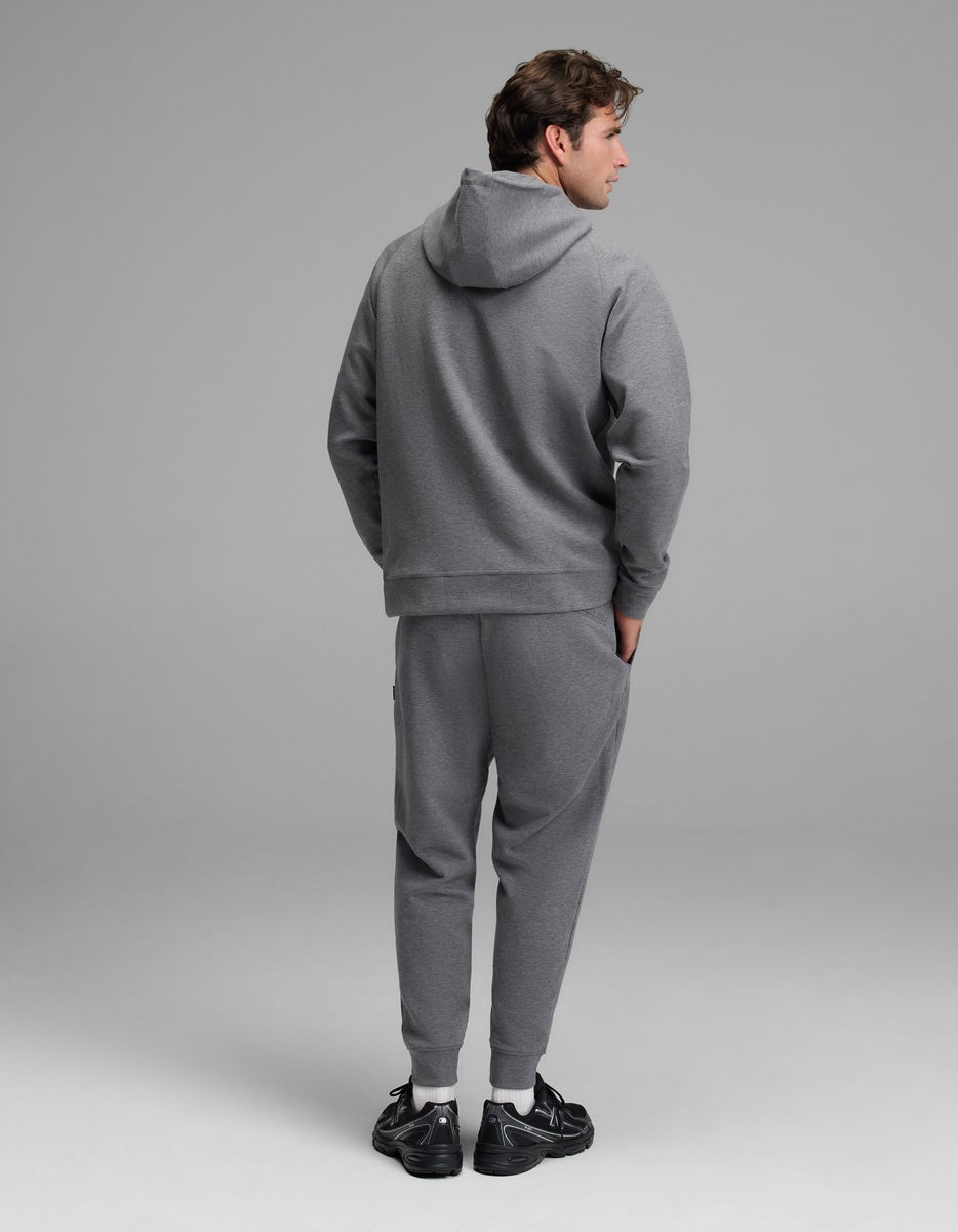 A person stands facing away, wearing the Cozy Earth Men’s StretchTech Hoodie & Jogger Set in gray with hands in pockets. The background is plain and neutral, and they are wearing black athletic sneakers.