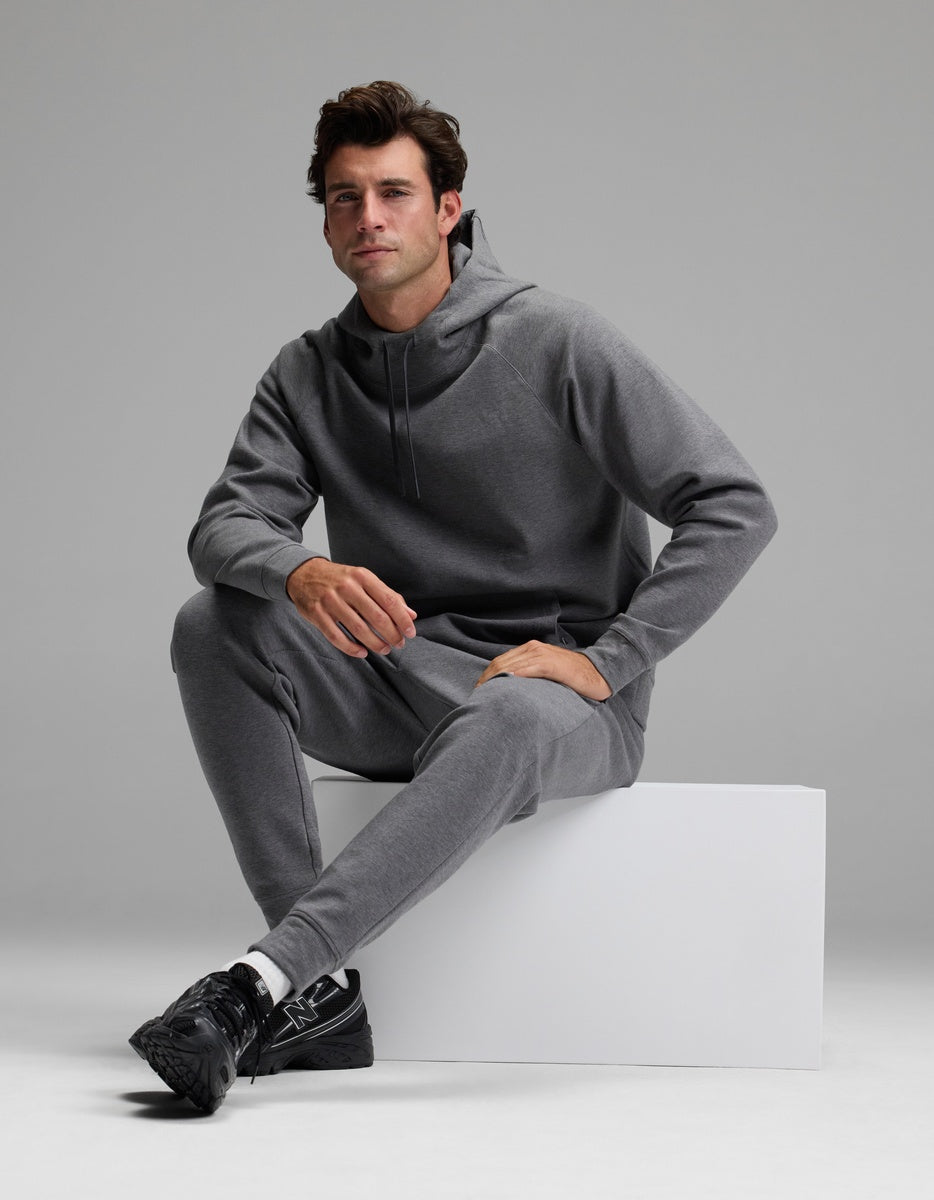A man models the Cozy Earth Men’s StretchTech Hoodie & Jogger Set in gray, sitting on a white block against a plain gray background. He looks forward with a neutral expression and pairs the outfit with black sneakers.