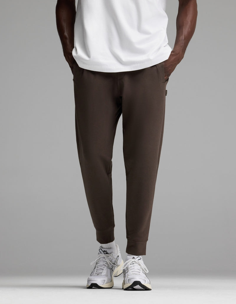 A person is shown from the waist down wearing Cozy Earth’s Men’s StretchTech Jogger in dark brown, paired with a white T-shirt and white sneakers, standing against a plain gray background.