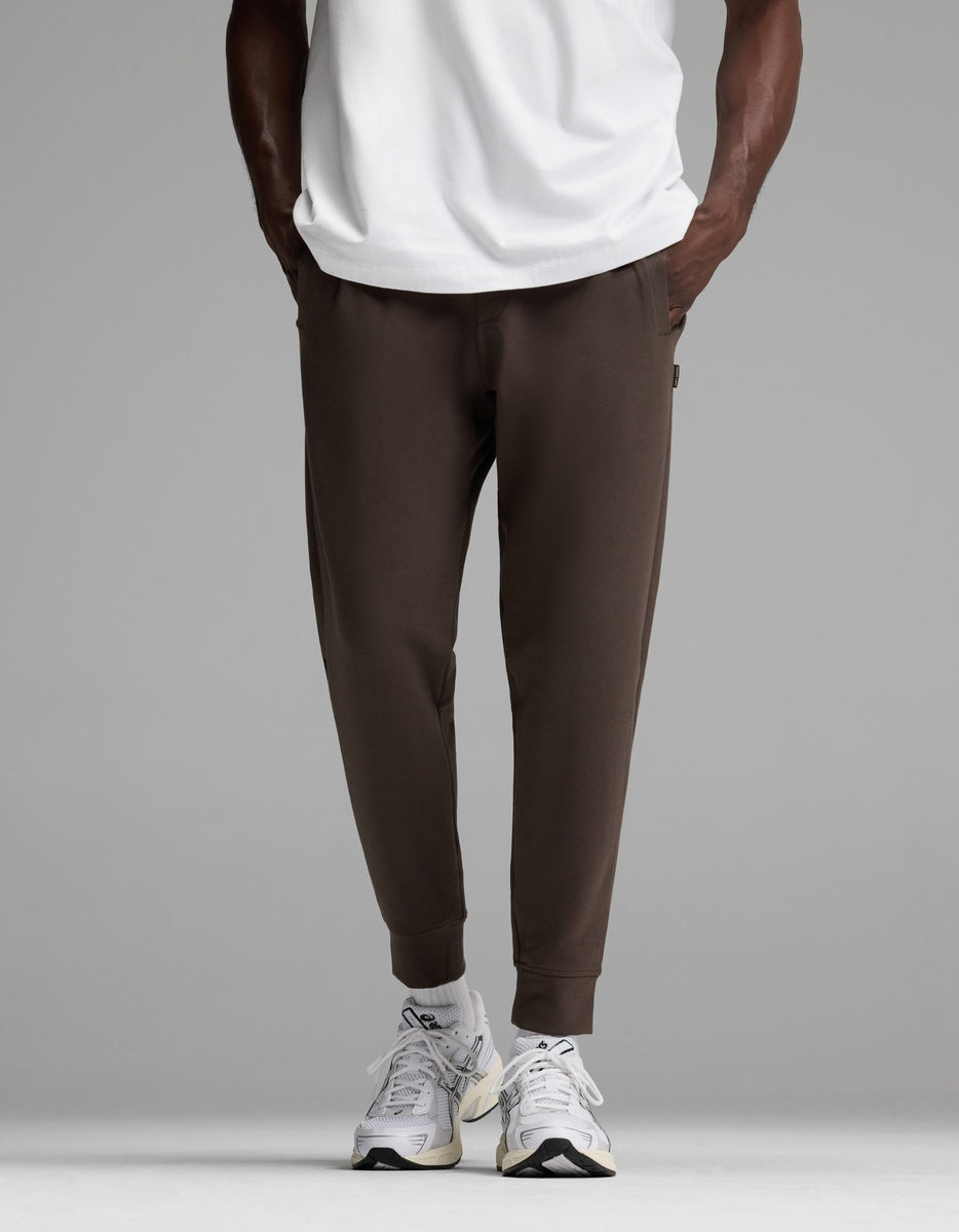 A person is shown from the waist down wearing Cozy Earth’s Men’s StretchTech Jogger in dark brown, paired with a white T-shirt and white sneakers, standing against a plain gray background.|Color:Cacao