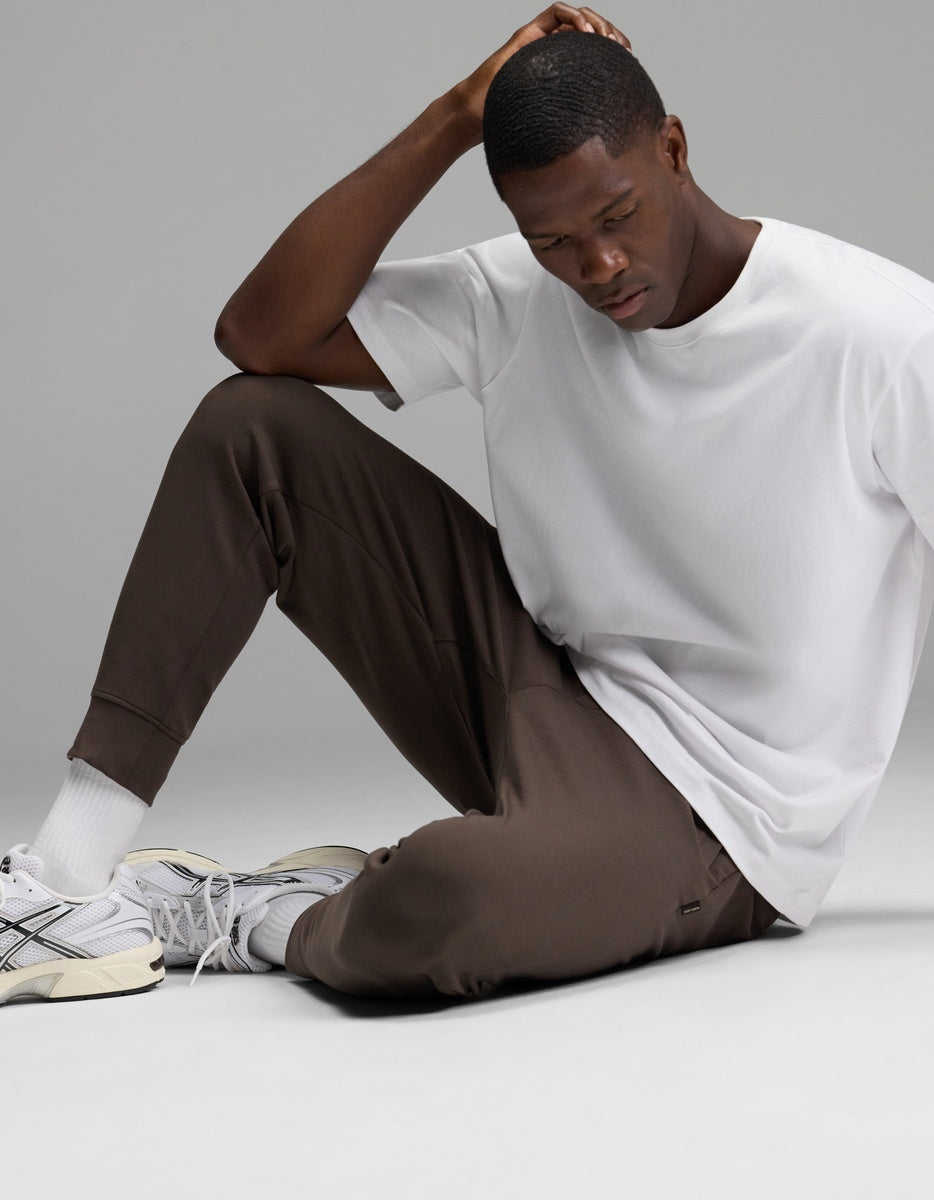 A man sits on the floor wearing a white t-shirt, white socks, gray sneakers, and Cozy Earth’s Men’s StretchTech Jogger in brown. He rests one hand on his head and looks down thoughtfully against a plain background.