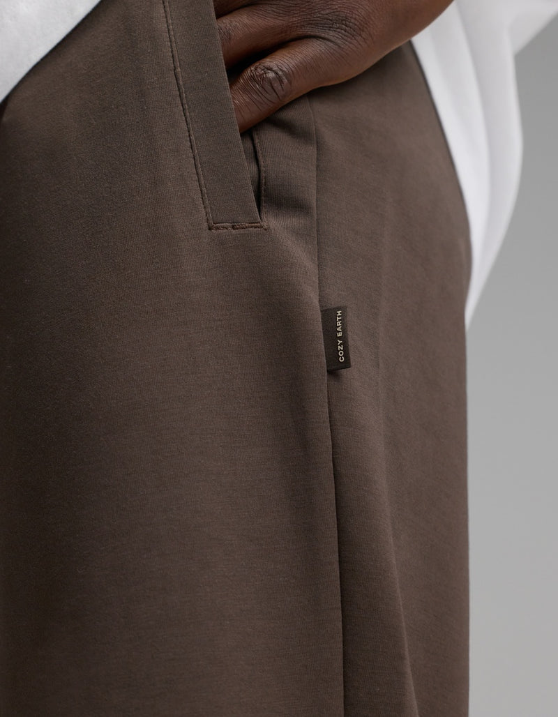 A close-up of a person’s hand in the pocket of Cozy Earth Men’s StretchTech Jogger pants in brown, featuring a small fabric tag with text. The person is also wearing a white shirt.