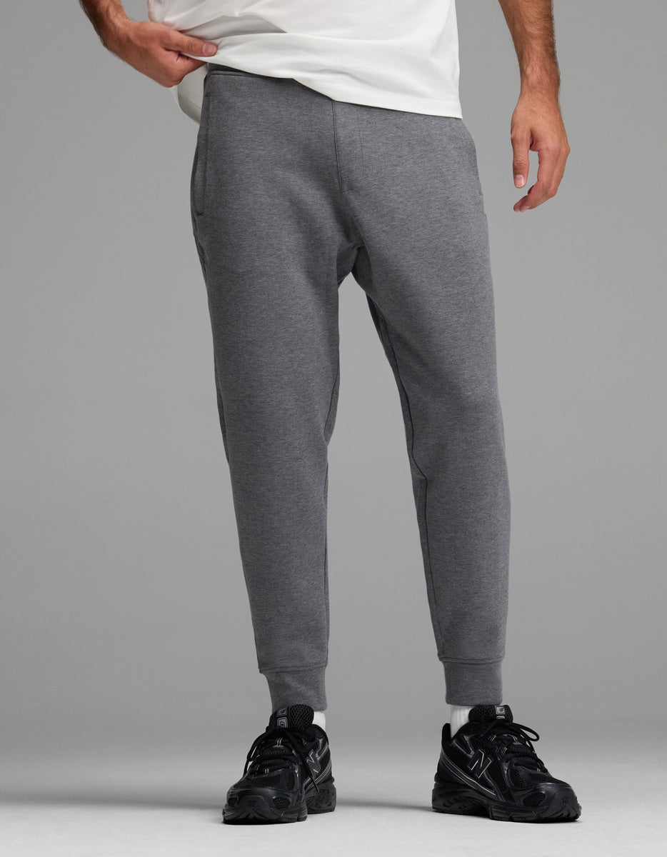 A person stands against a plain gray background, wearing Cozy Earth Men’s StretchTech Jogger sweatpants in gray, paired with black sneakers and a white shirt. Only the lower half of the body is visible.|Color:Coal Heather
