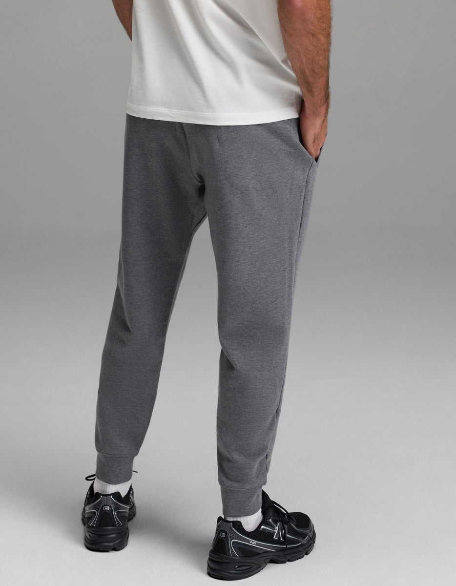 A person stands with hands in pockets, wearing a white t-shirt, Cozy Earth Men’s StretchTech Jogger in gray, and black athletic shoes. The photo is taken from behind, highlighting the legs and lower back.