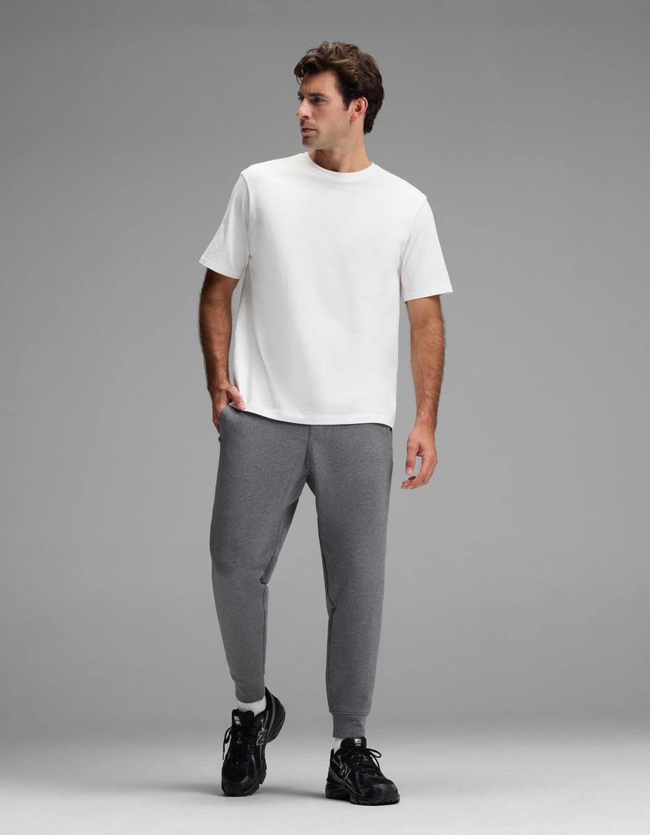 A man wearing a plain white T-shirt, Cozy Earth Men’s StretchTech Jogger pants in gray, and black sneakers stands against a plain gray background, looking to his right.