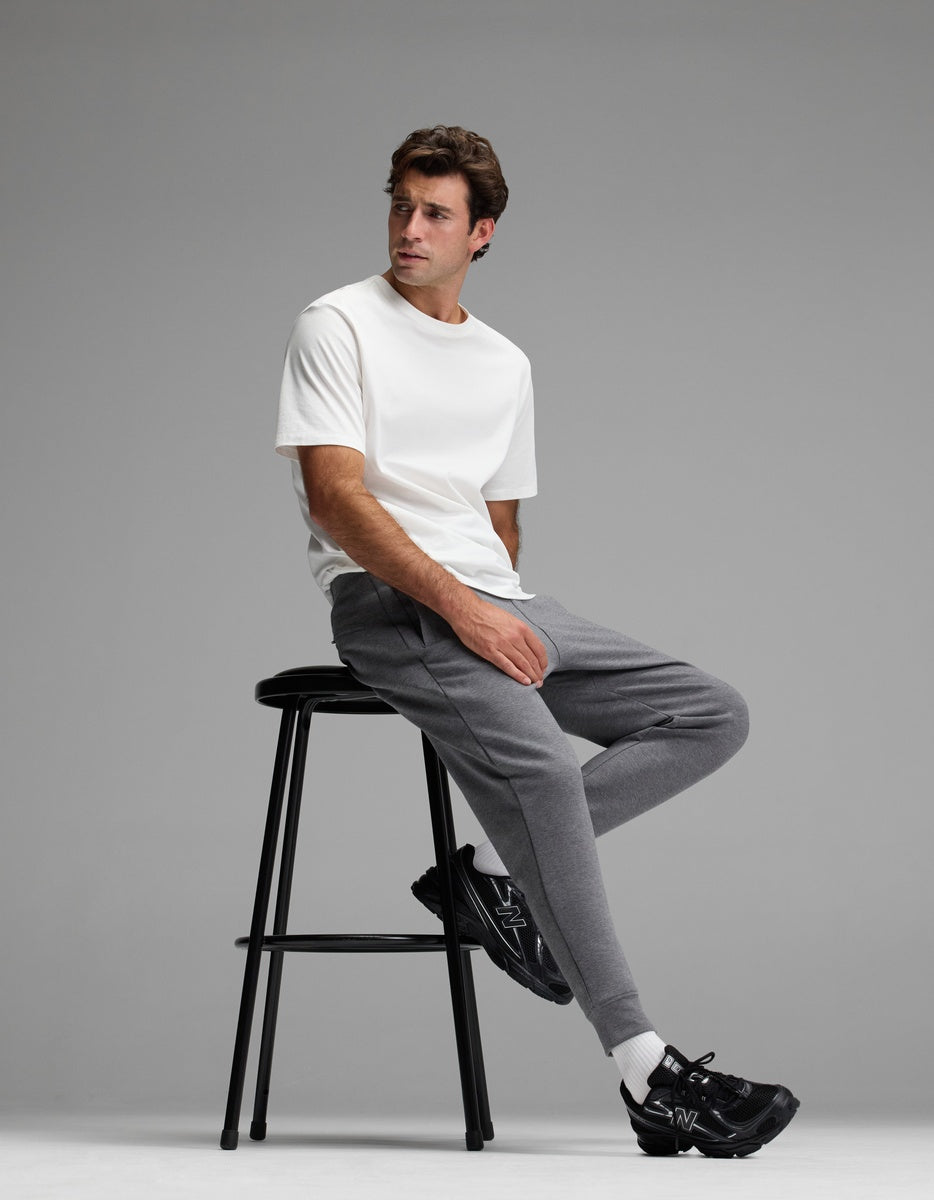 A man wearing a white t-shirt, white socks, black sneakers, and Cozy Earth Men’s StretchTech Jogger sits on a tall black stool against a plain gray background.