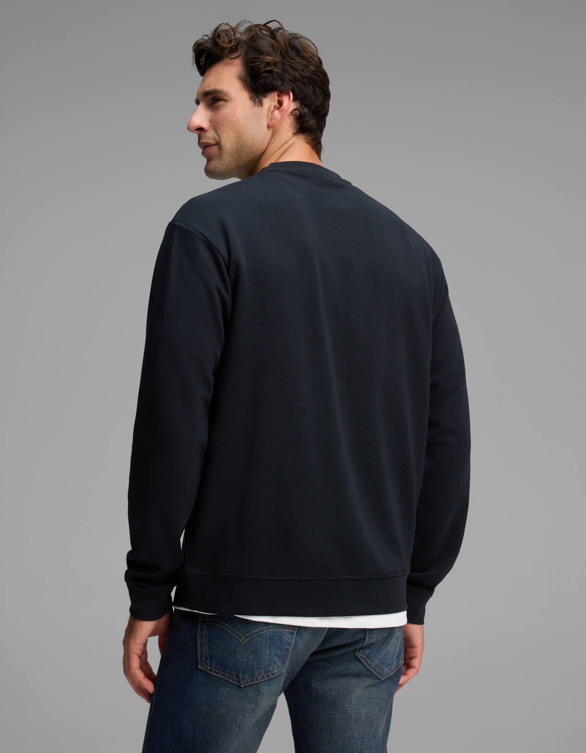 A man with dark hair models the Cozy Earth Men's Versa Crewneck in black with blue jeans, standing with his back to the camera and looking over his left shoulder against a plain gray background.