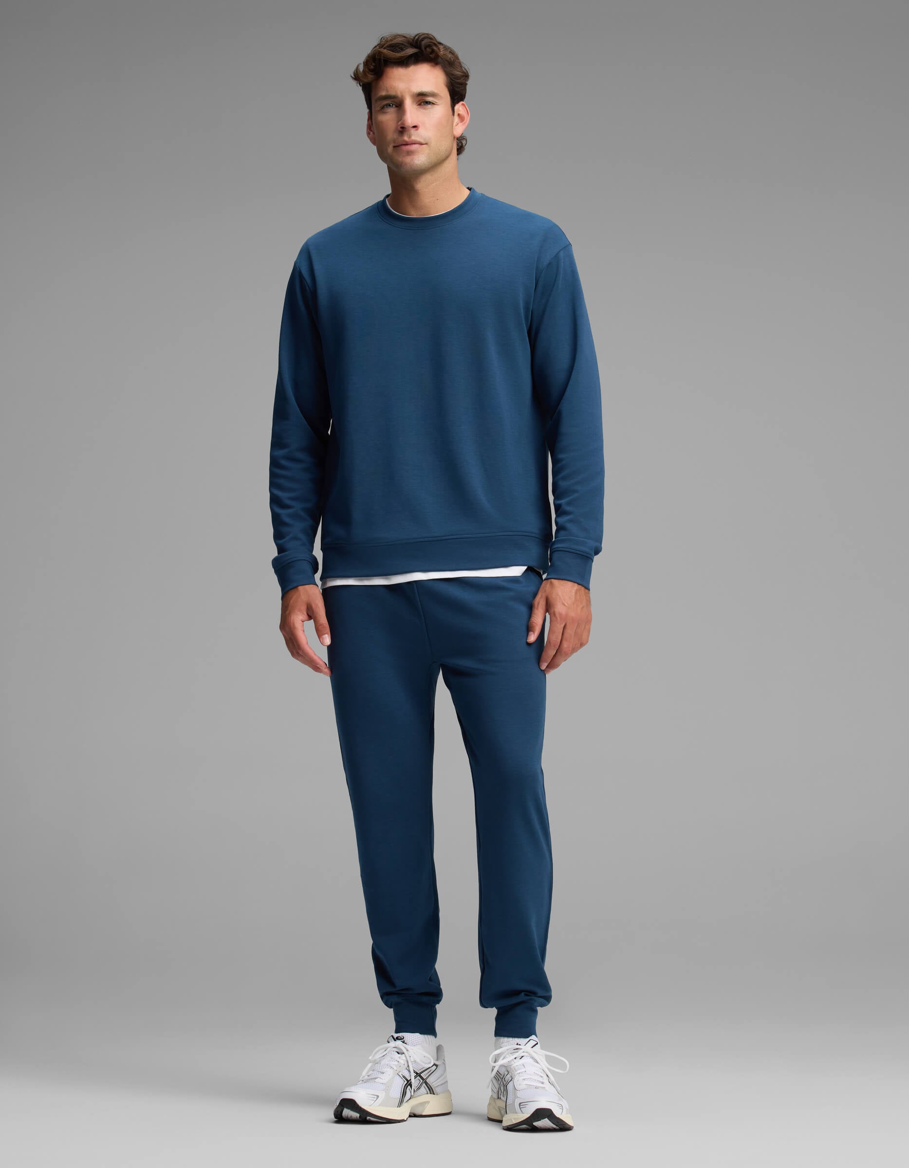 A man stands facing forward against a plain gray background, wearing the Cozy Earth Men's Versa Crewneck & Jogger Set in blue, paired with white sneakers and a white shirt visible beneath the sweatshirt.