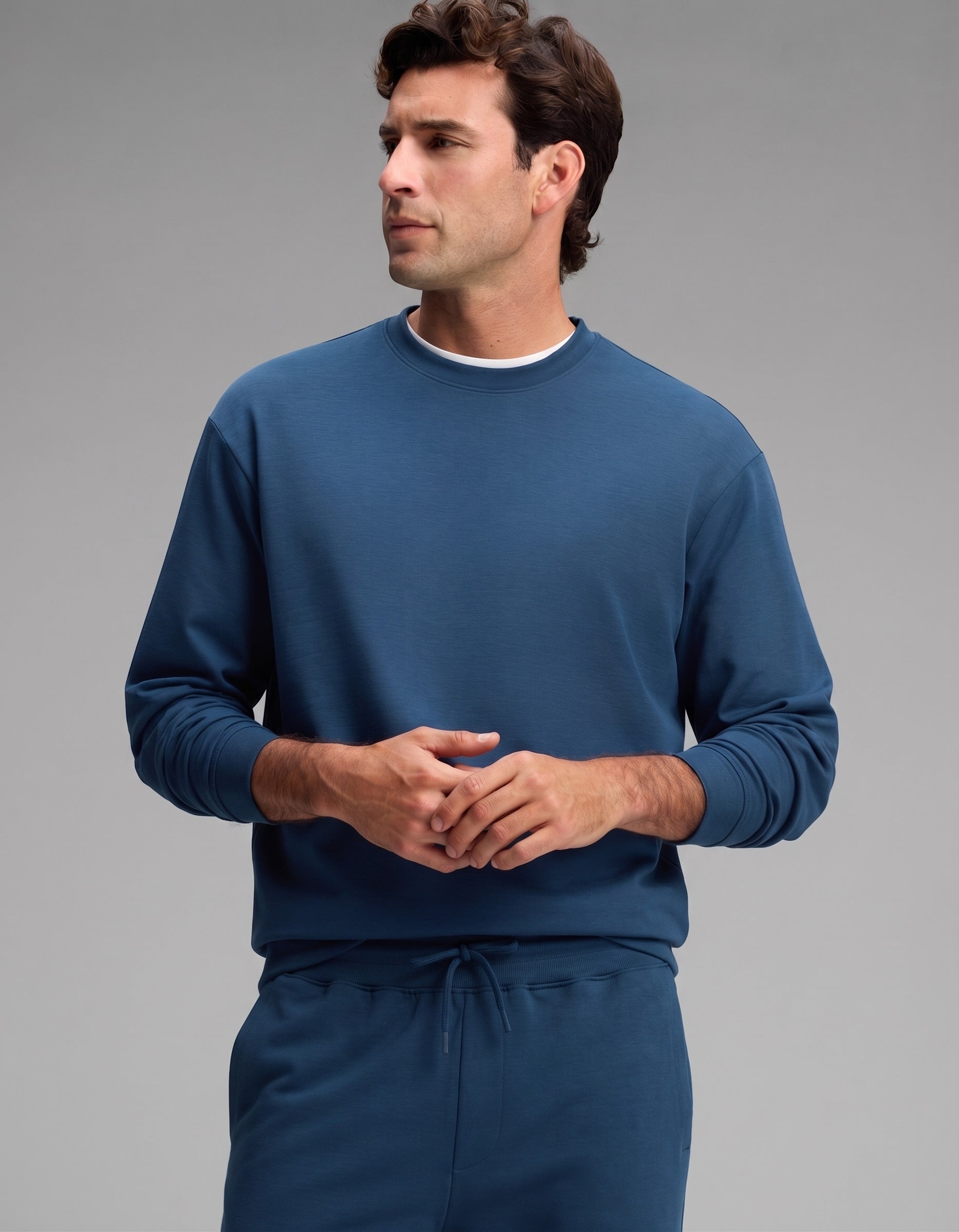 A man models the Cozy Earth Men's Versa Crewneck & Jogger Set in blue, standing against a plain gray background with his hands lightly clasped and looking to the left.