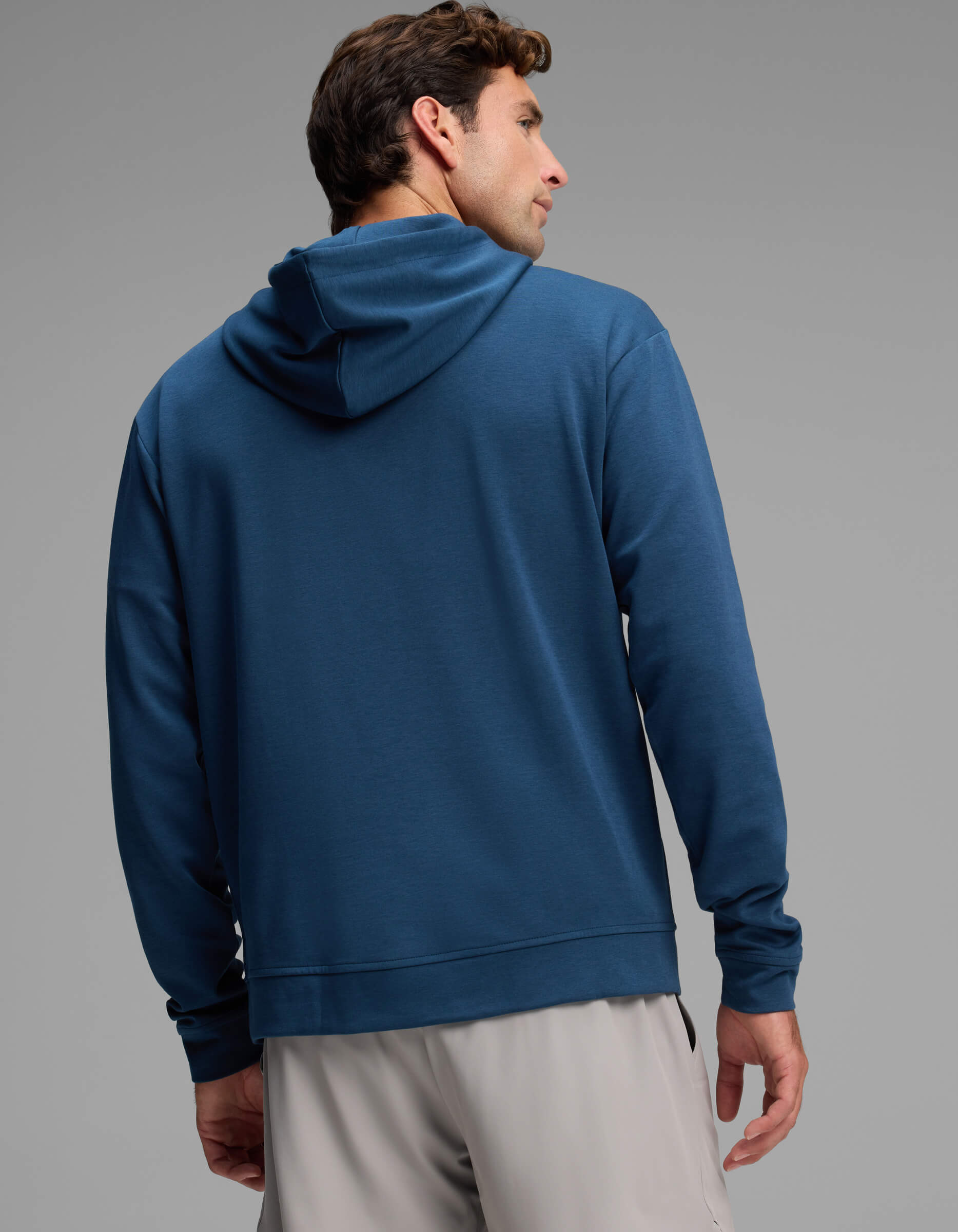 A man wearing the Cozy Earth Men's Versa Hoodie in blue and light gray shorts stands facing away, glancing over his left shoulder against a plain gray background.|Color:Obsidian