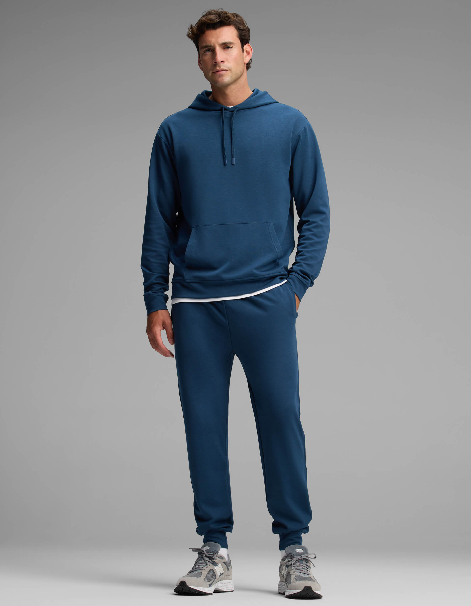A man stands against a gray background wearing the Cozy Earth Men's Versa Hoodie & Jogger Set in blue, paired with a white T-shirt and gray sneakers. He looks forward with a neutral expression, one hand by his side and the other slightly bent.|Color:Obsidian