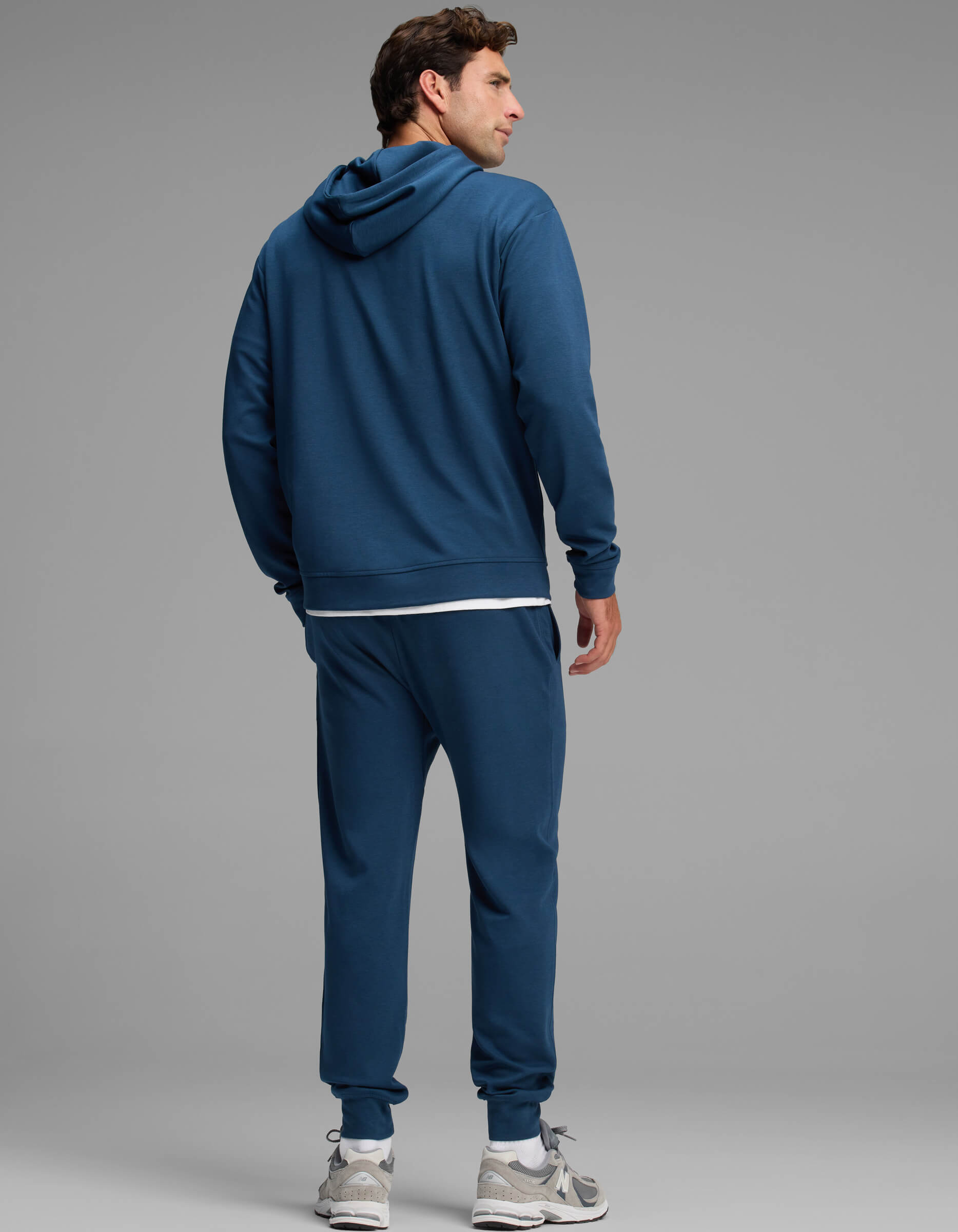A man stands facing away, wearing the Cozy Earth Men's Versa Hoodie & Jogger Set in blue, paired with white sneakers against a plain gray background.|Color:Obsidian