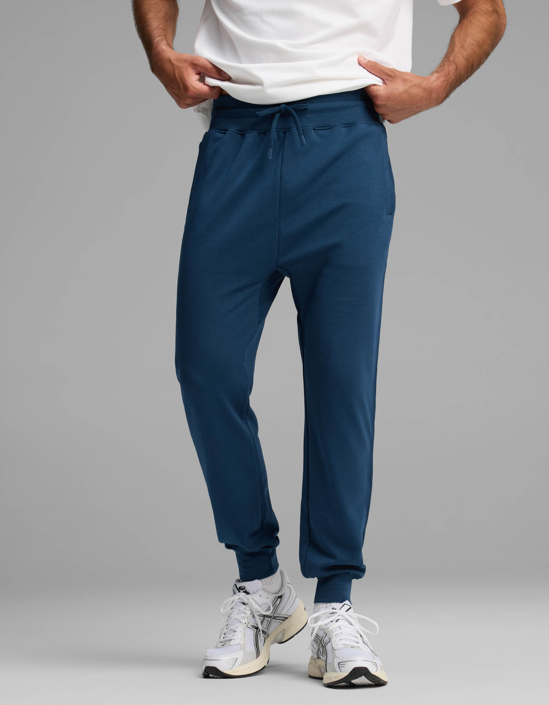 A person wearing a white shirt, Cozy Earth Men's Versa Jogger in blue, and white athletic shoes stands against a plain gray background with their hands in their pockets.