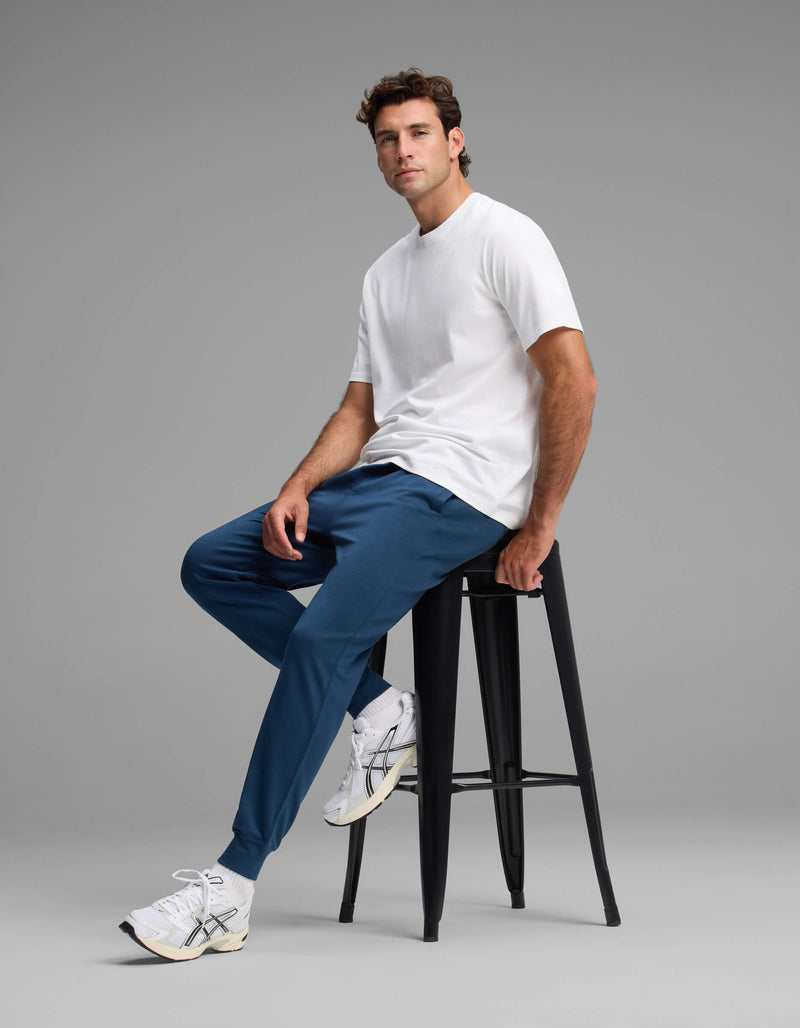 A man wearing the Cozy Earth Men's Versa Jogger in blue and a white t-shirt sits on a tall black stool against a plain gray background, paired with white sneakers and gazing slightly to the side.
