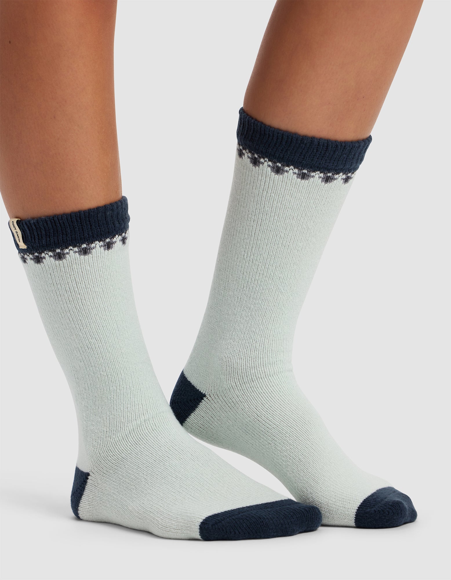 Two feet wearing Cozy Earth's The Mistletoe Plush Lounge Sock in light grey with dark navy-blue cuffs, toes, and heels. The socks feature a small dotted pattern near the top and a branded tag on one cuff. Background is plain white.|Color:Mist