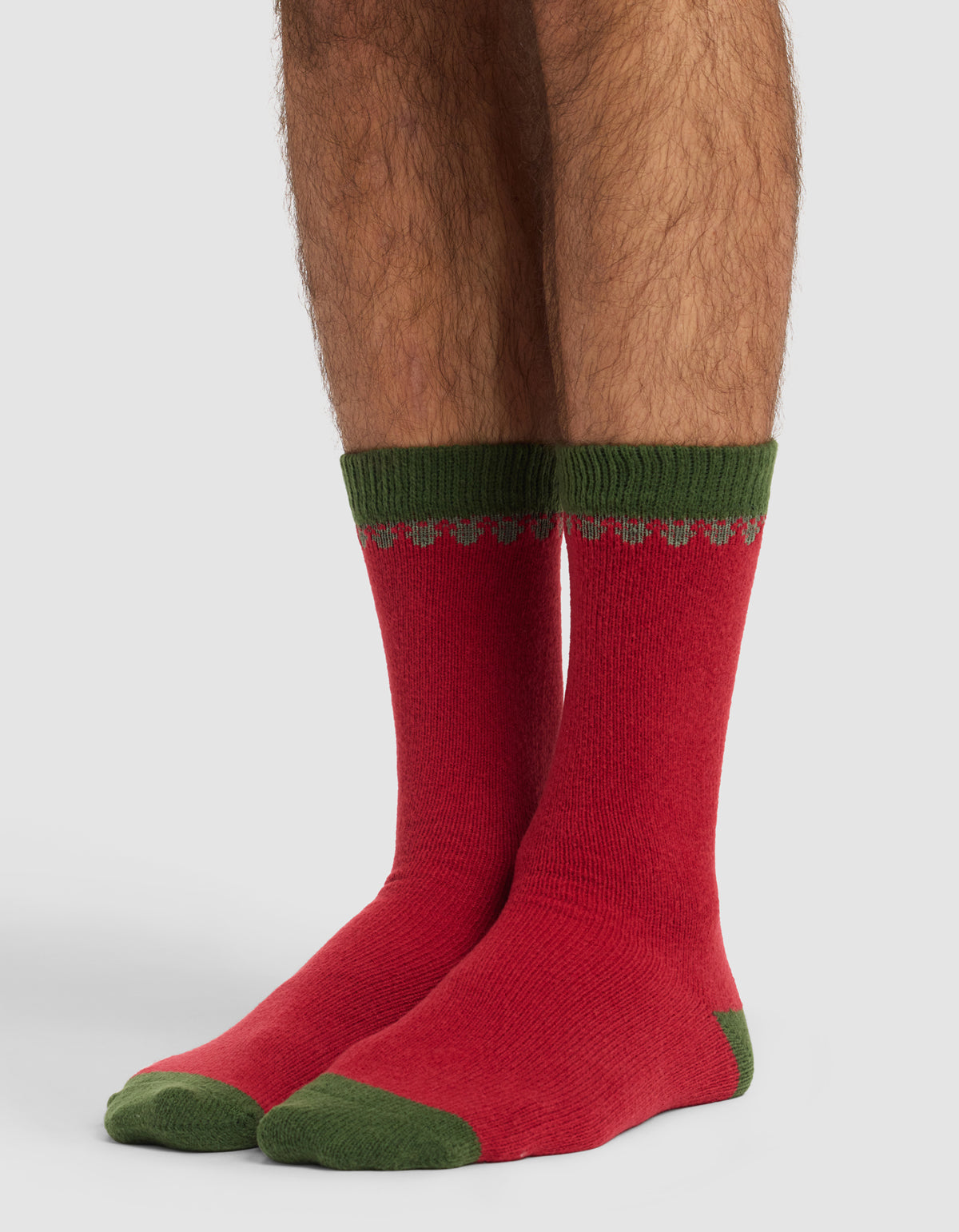 A person wears The Mistletoe Plush Lounge Sock by Cozy Earth—red socks with green heels, toes, and cuffs plus a small green and gray pattern near the top. The plain background is light-colored.|Color:Cranberry