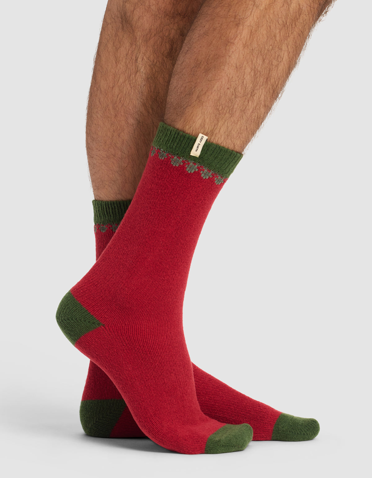 A person wearing The Mistletoe Plush Lounge Sock by Cozy Earth—red with green toes, heels, and cuffs, featuring a small decorative tag and a subtle zigzag pattern near the top—stands against a plain light gray background.|Color:Cranberry