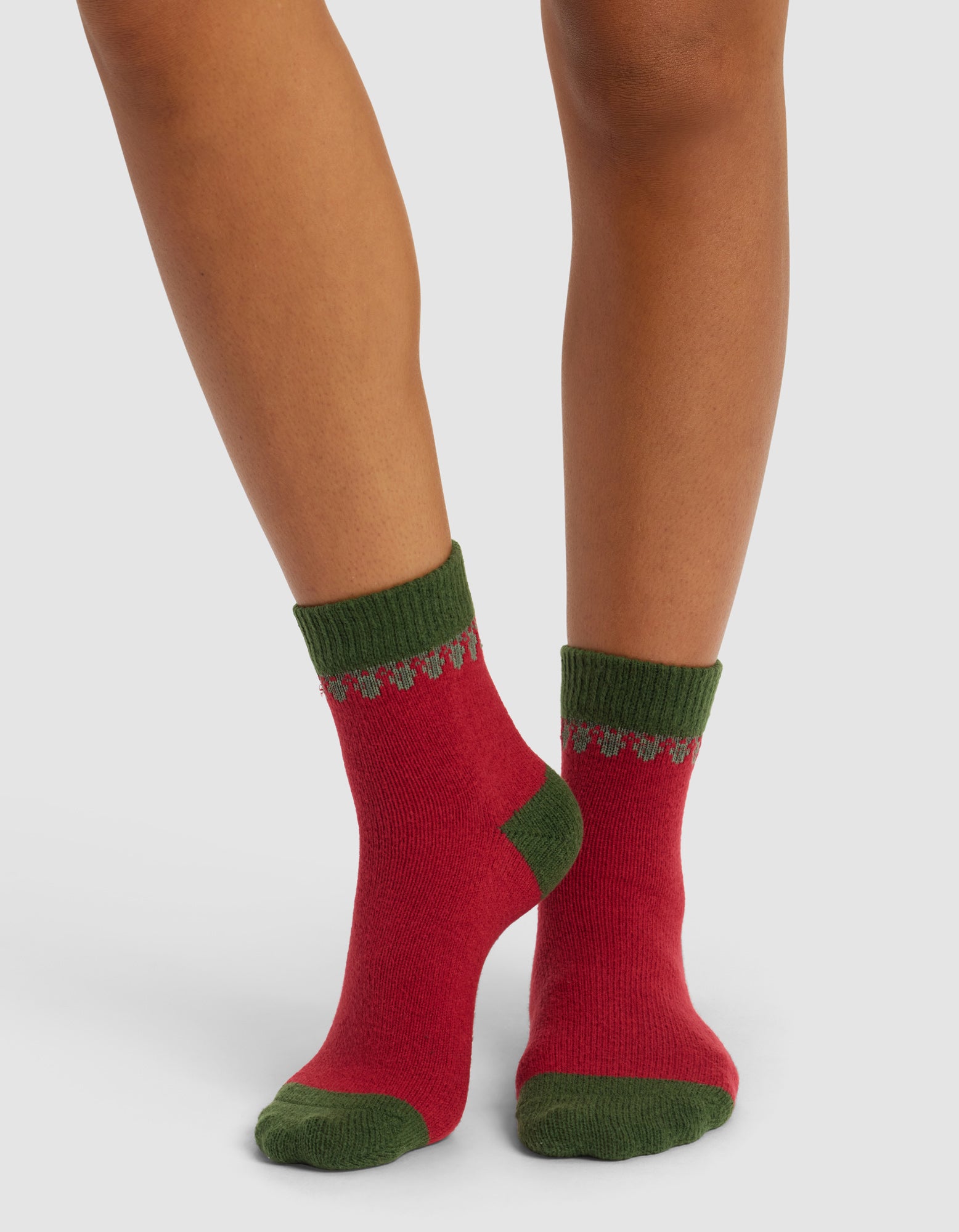 A person wearing The Mistletoe Plush Modern Crew Sock by Cozy Earth, featuring red with green heels, toes, and cuffs accented by a green and gray decorative pattern. Background is plain white.|Color:Cranberry