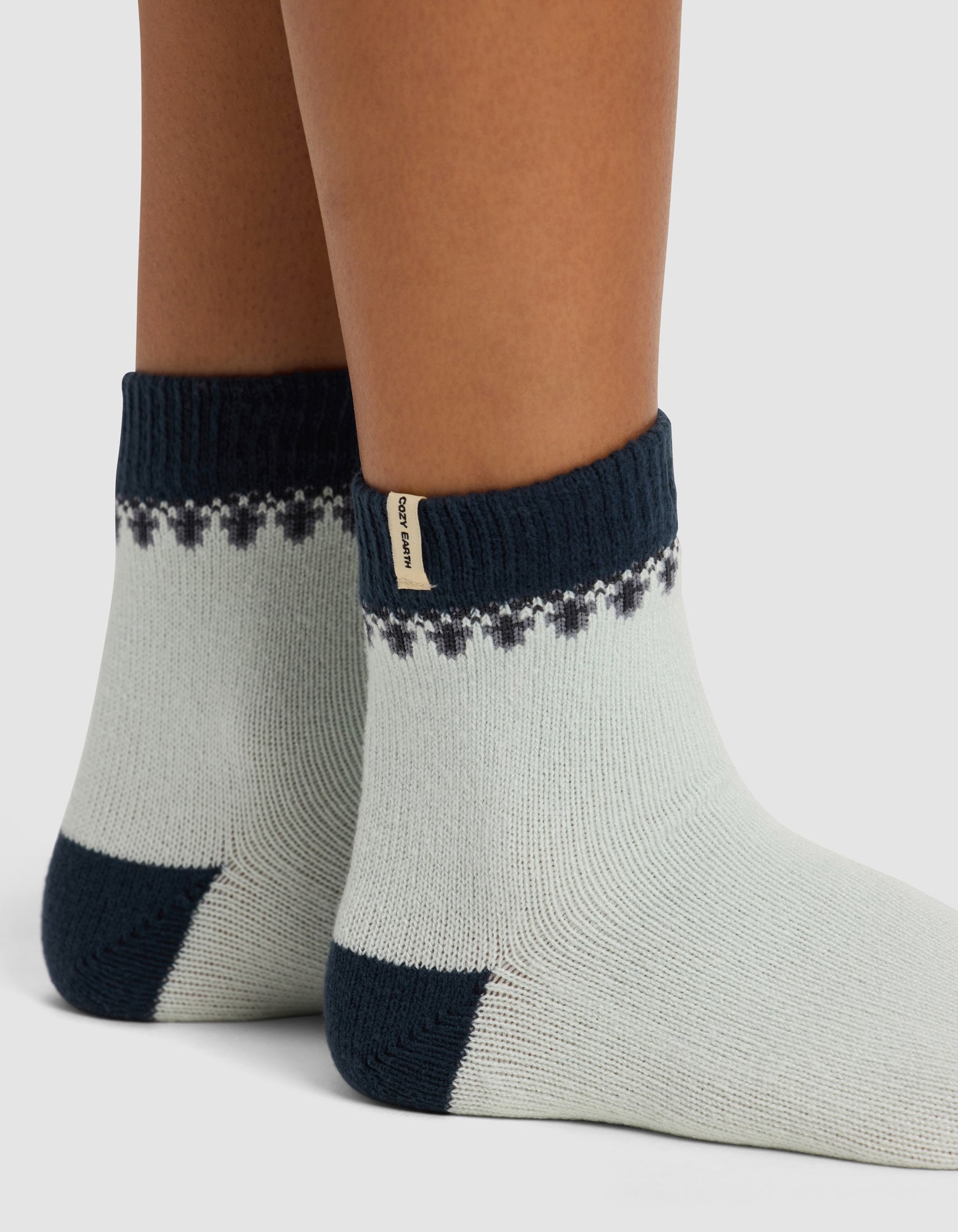 Close-up of two feet wearing Cozy Earth’s The Mistletoe Plush Modern Crew Socks—thick, light-colored with dark heels, toes, and cuffs, plus a pattern near the top and a small beige tag on one cuff.
