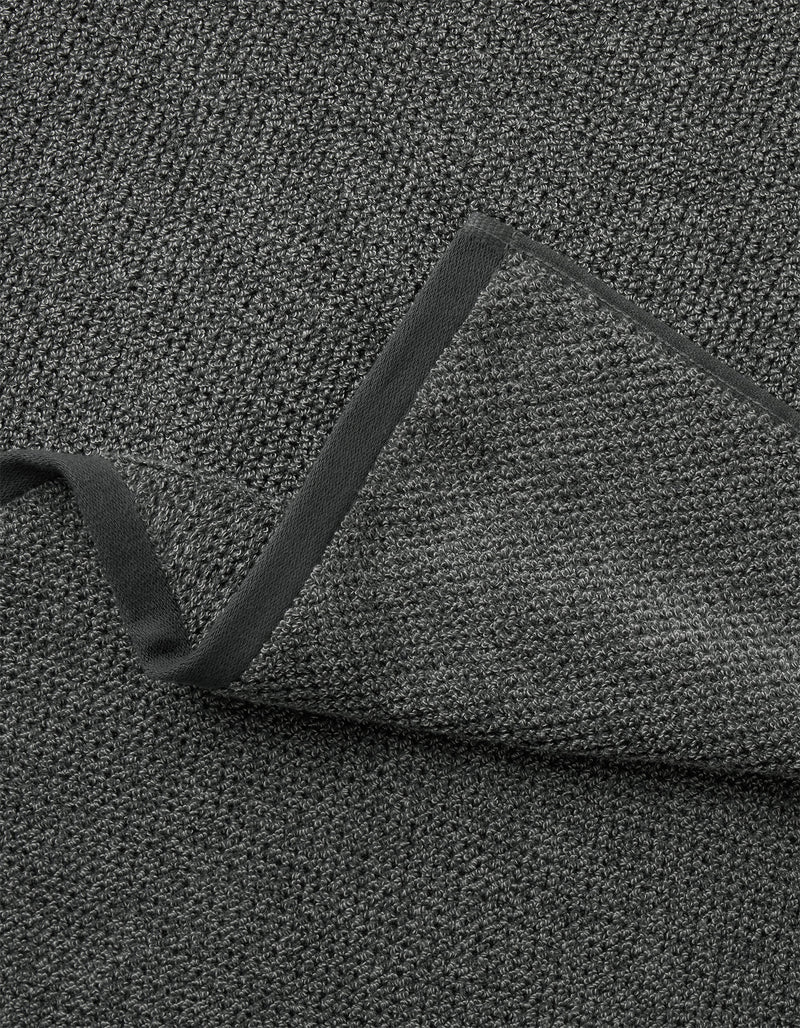 A close-up of Cozy Earth's Nantucket Bath Towel Set shows its textured dark gray fabric with a folded corner, revealing the same woven material on both sides and a smooth, dark border along the edge.