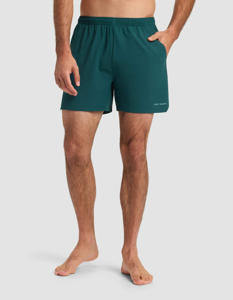 A barefoot man stands against a plain background, wearing Cozy Earth Men's Day & Night Short 5" in teal with an elastic waistband and one hand in his pocket.