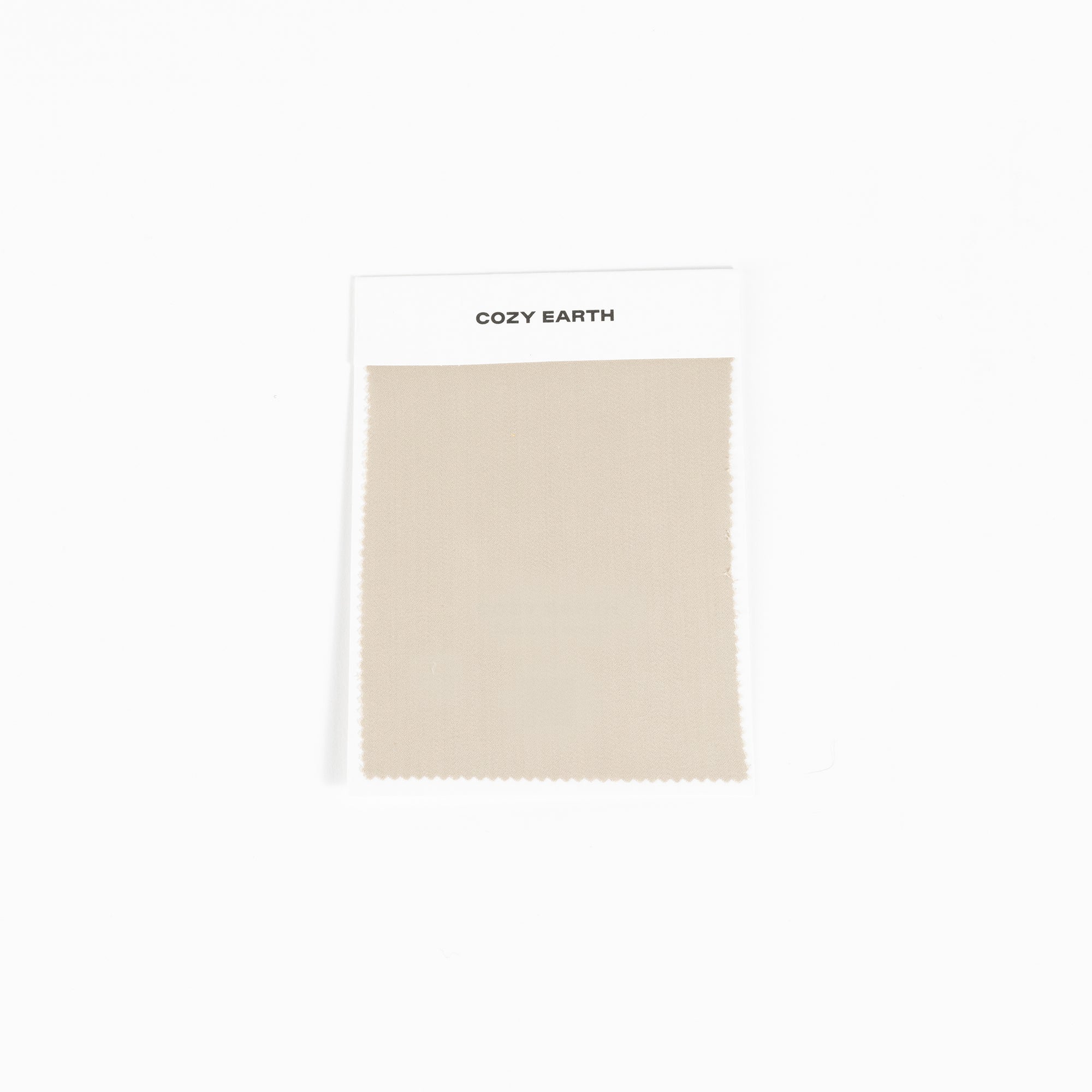 A square fabric sample from Cozy Earth, identified as the Bamboo Sheet Swatch. The swatch is a simple beige color with a zigzag edge design and is attached to a white card. 