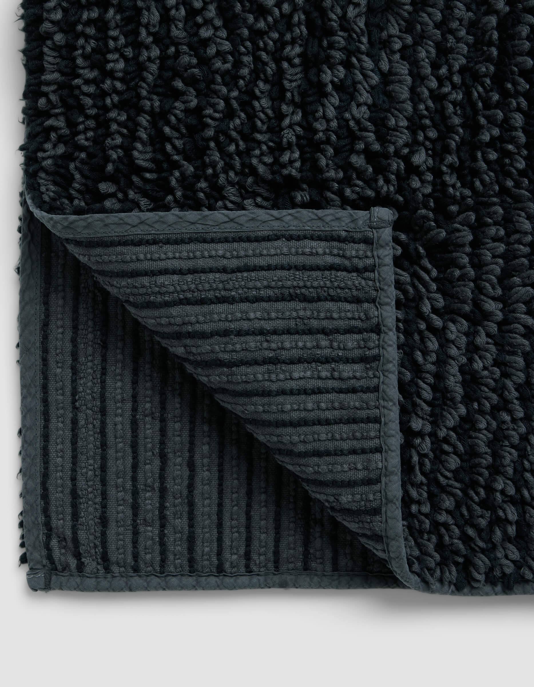 Close-up of the Cozy Earth Plush Loop Bath Mat with a corner folded, highlighting its ribbed, non-slip underside and thick, soft plush pile on top.|Color:Graphite