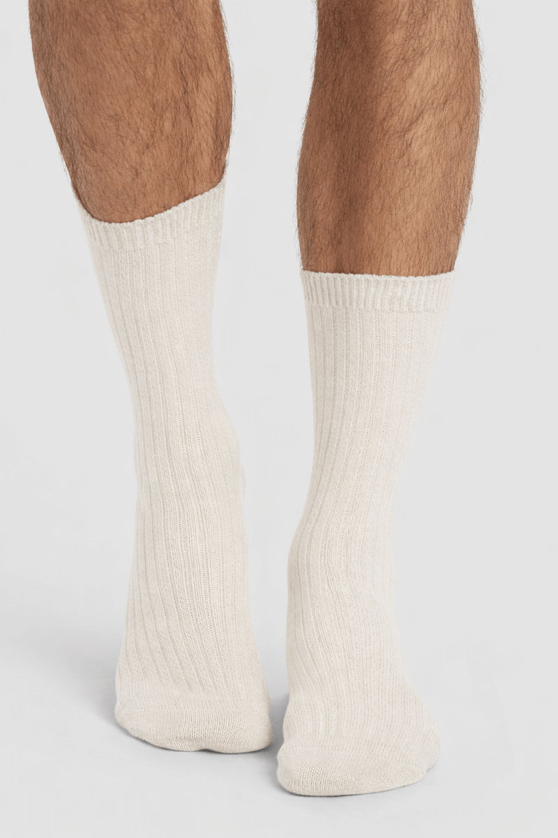 A close-up of a person's lower legs wearing Cozy Earth's Men's Plush Lounge Calf Sock 3-Pack in cream, ribbed design, standing on a white surface.