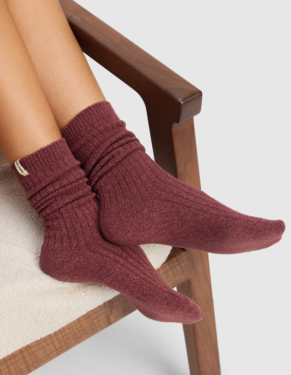 A person sits on a cushioned wooden chair with crossed legs, wearing Cozy Earth’s Plush Lounge Calf Socks in maroon from the 3-Pack. The textured socks are slightly slouched. The background is plain and light-colored.|Color:Burgundy/Rouge/Dusty Rose
