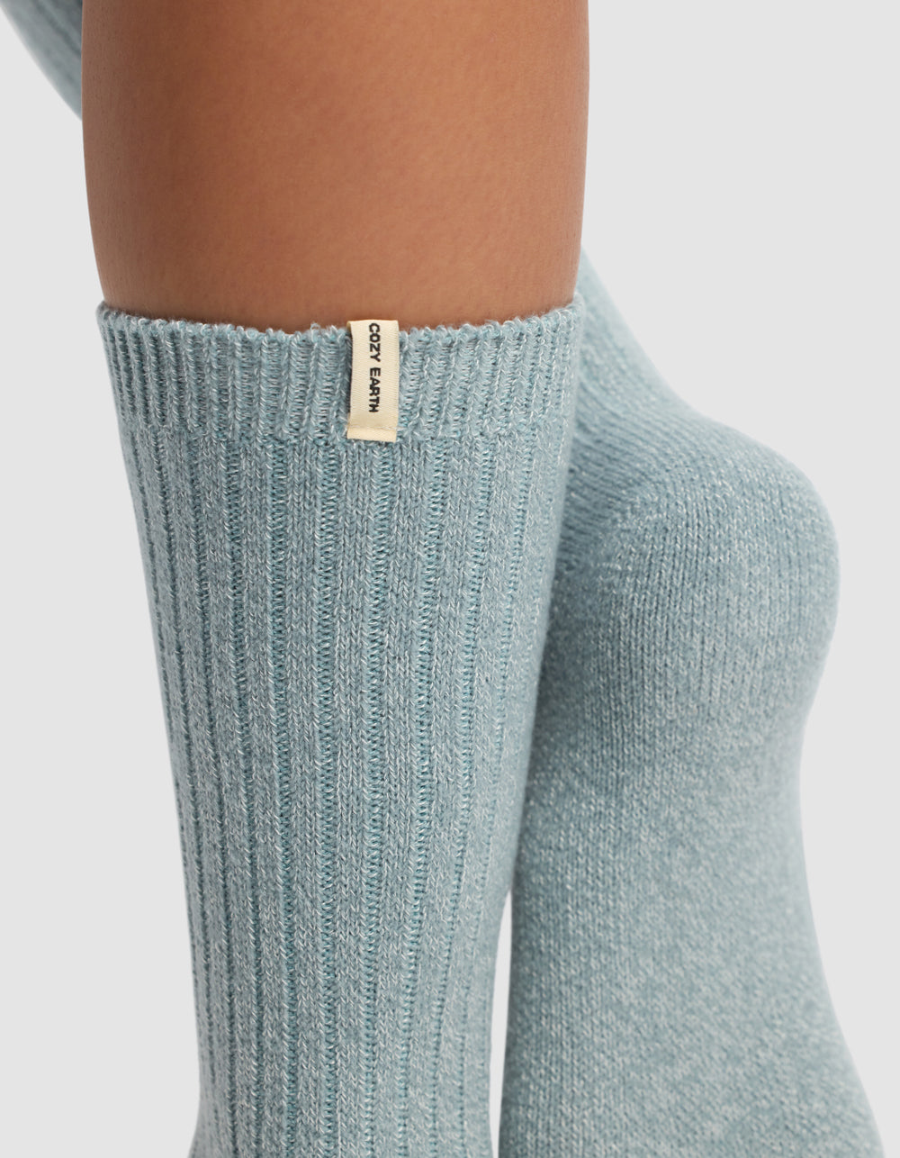 Close-up of lower legs wearing light blue ribbed socks from Cozy Earth's The Plush Lounge Calf Sock 3-Pack, featuring a small beige tag with the brand name. Background is plain white.
